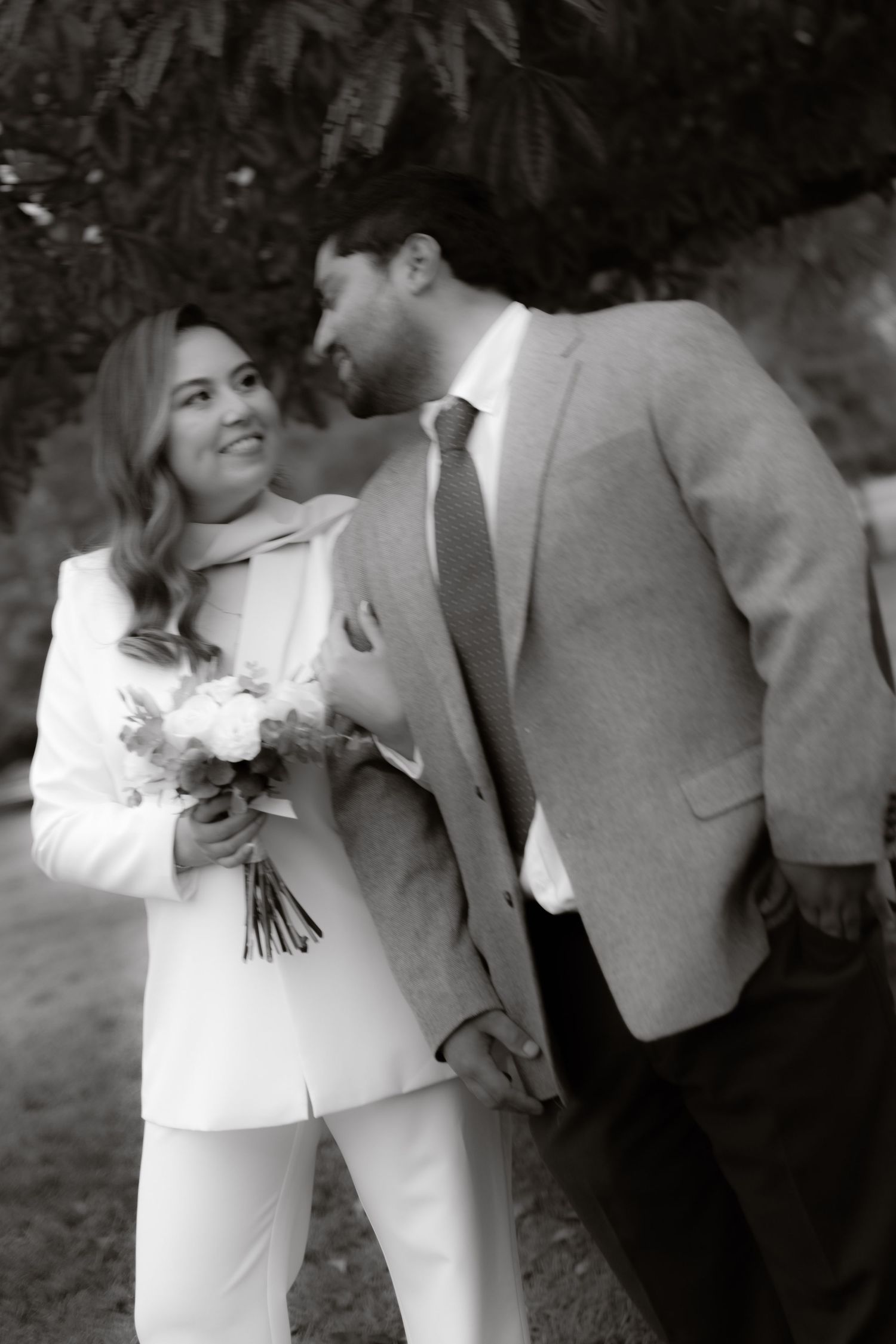 Black and white photo of smiling bride in white suit holding bouquet with groom in park.
