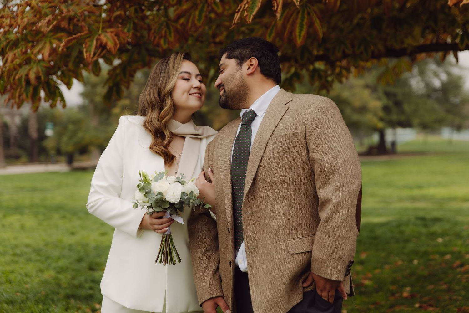 Bride in white blazer and groom in tan suit share a smile under colorful autumn leaves.