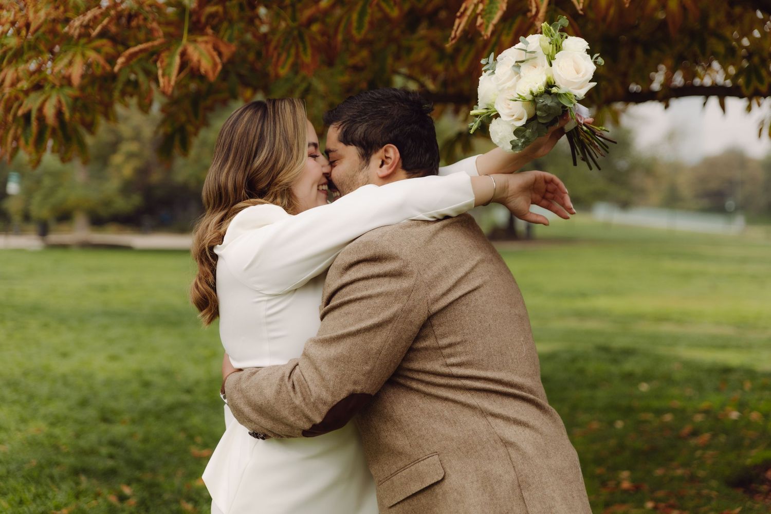 Newlyweds share a romantic kiss under a red autumn tree, bride holding white bouquet aloft.
