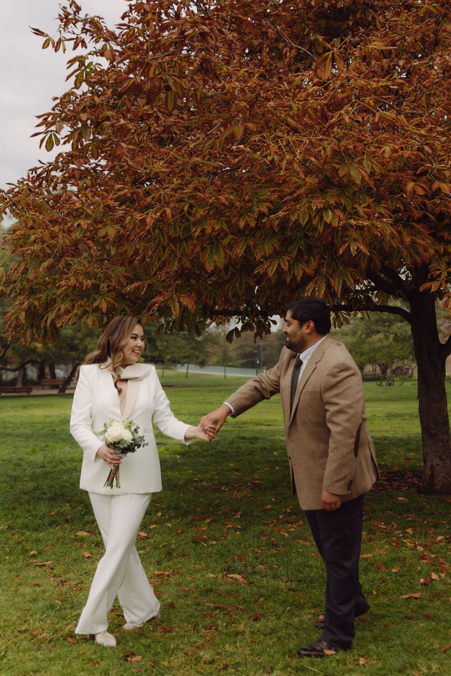 Bride and groom hold hands walking in park with stunning red autumn tree in background.