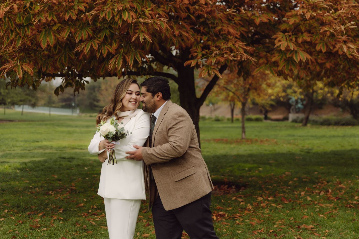 Bride in white suit holding bouquet embraced by groom in tan blazer under autumn tree in park.