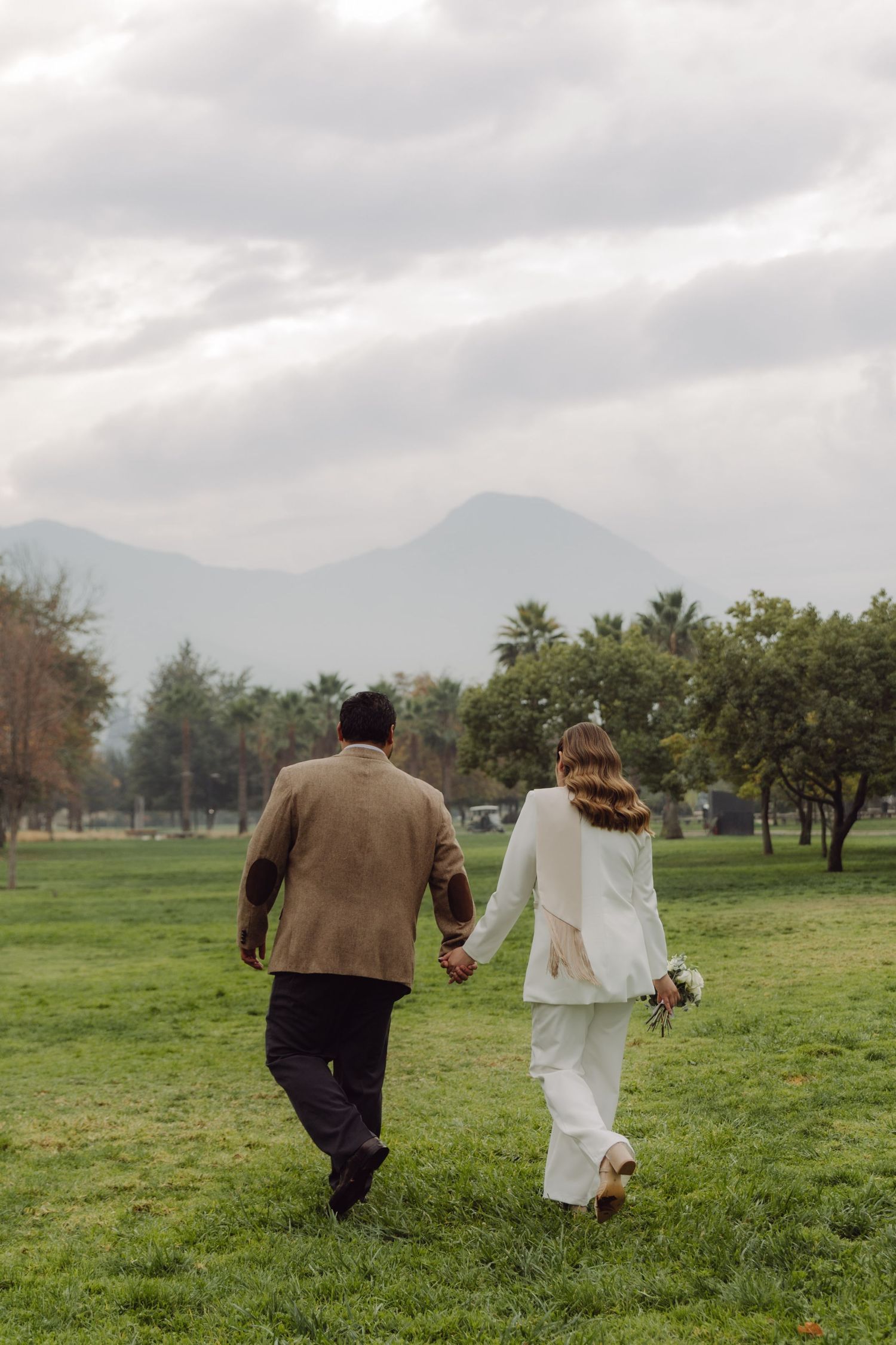 Newlywed couple walking hand in hand across green field with mountains and cloudy sky behind.