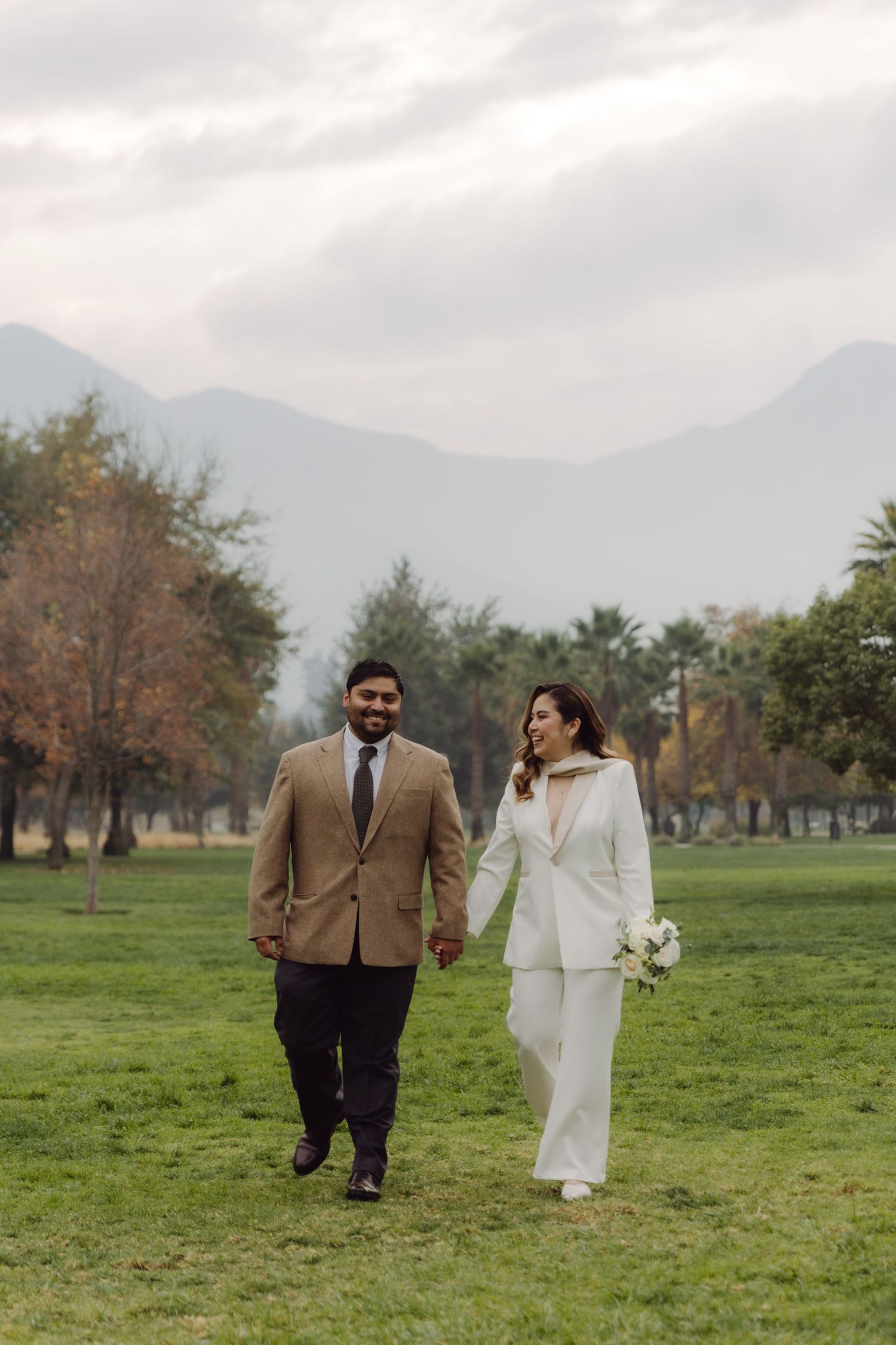 Smiling couple in wedding attire walking through park with mountain backdrop on overcast day.