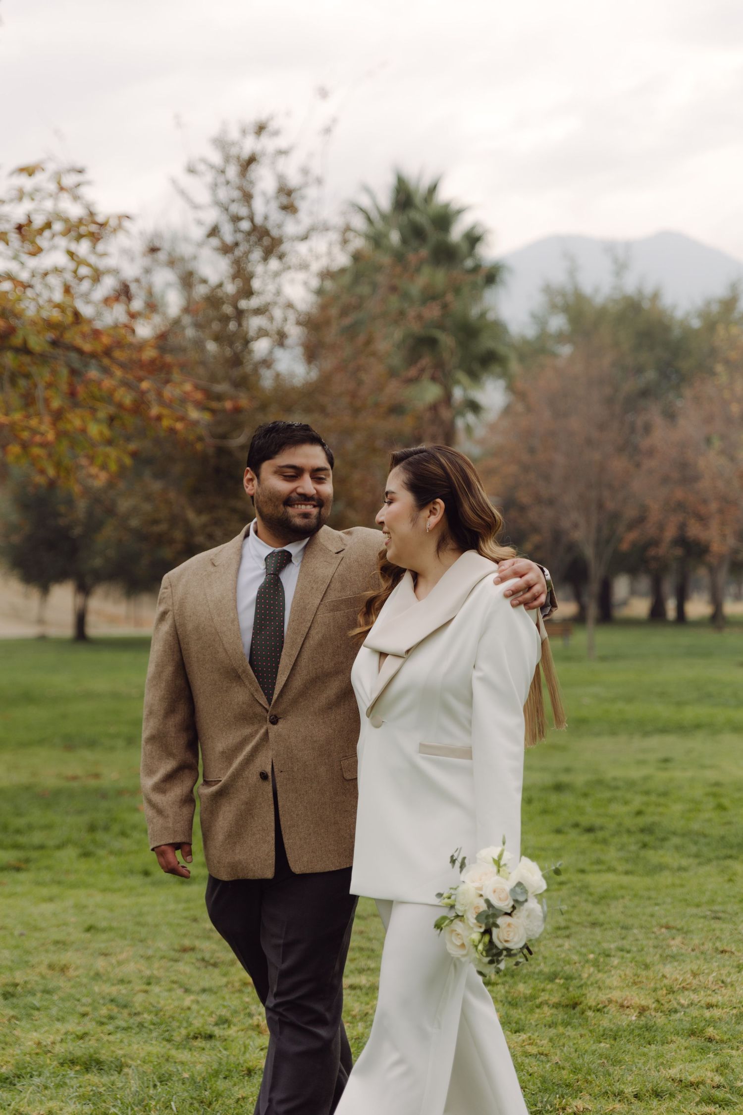 Groom in tan suit embracing bride in white blazer while strolling through autumn park setting.