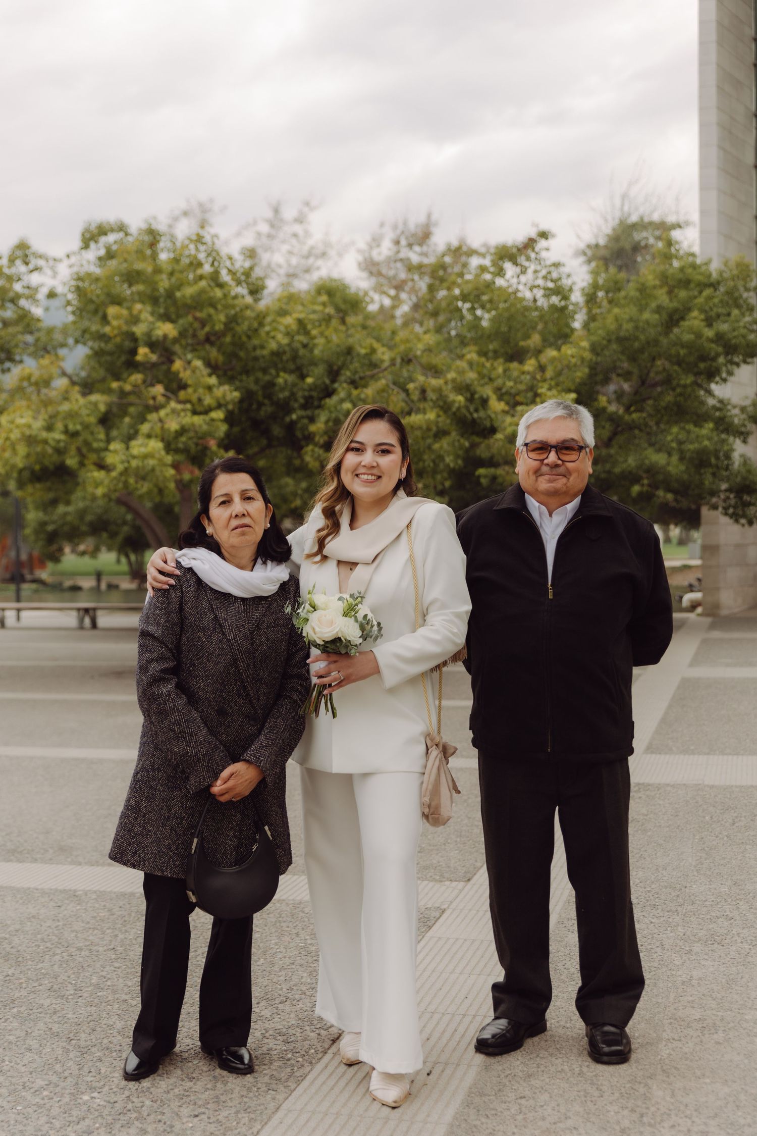Bride in white suit holding bouquet posing with elderly parents outside modern building.