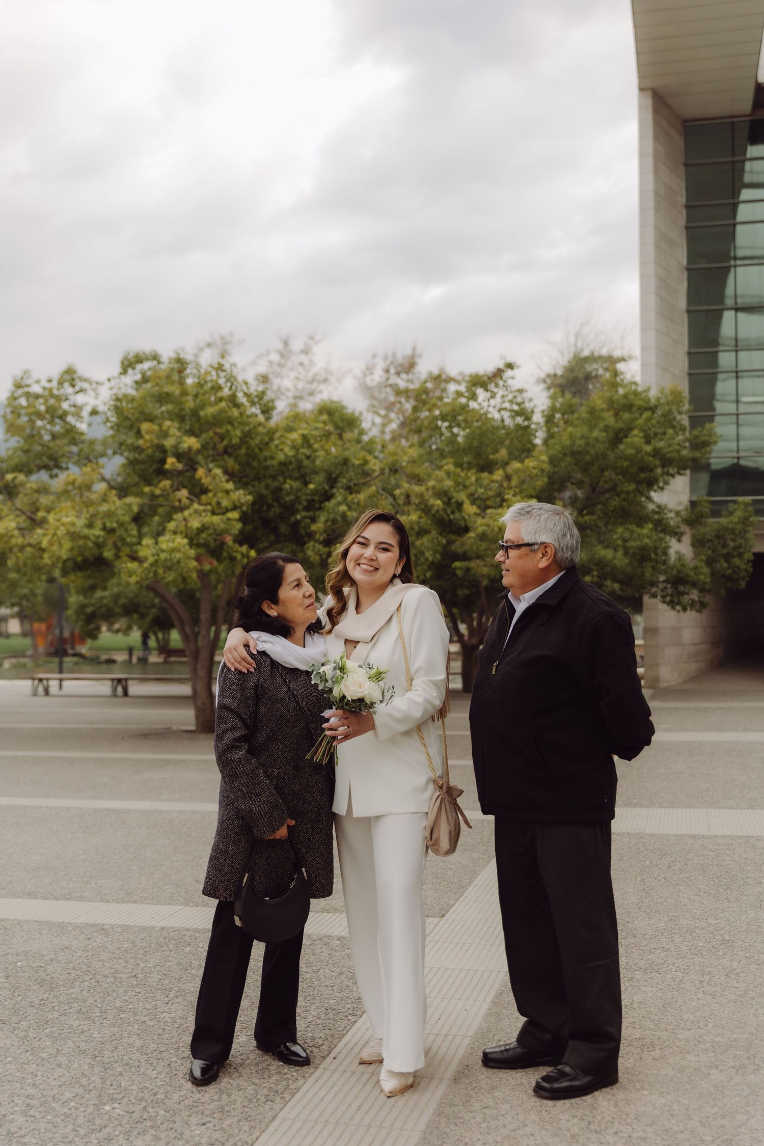 Bride in white wedding suit laughing with her parents outdoors near a modern glass building.