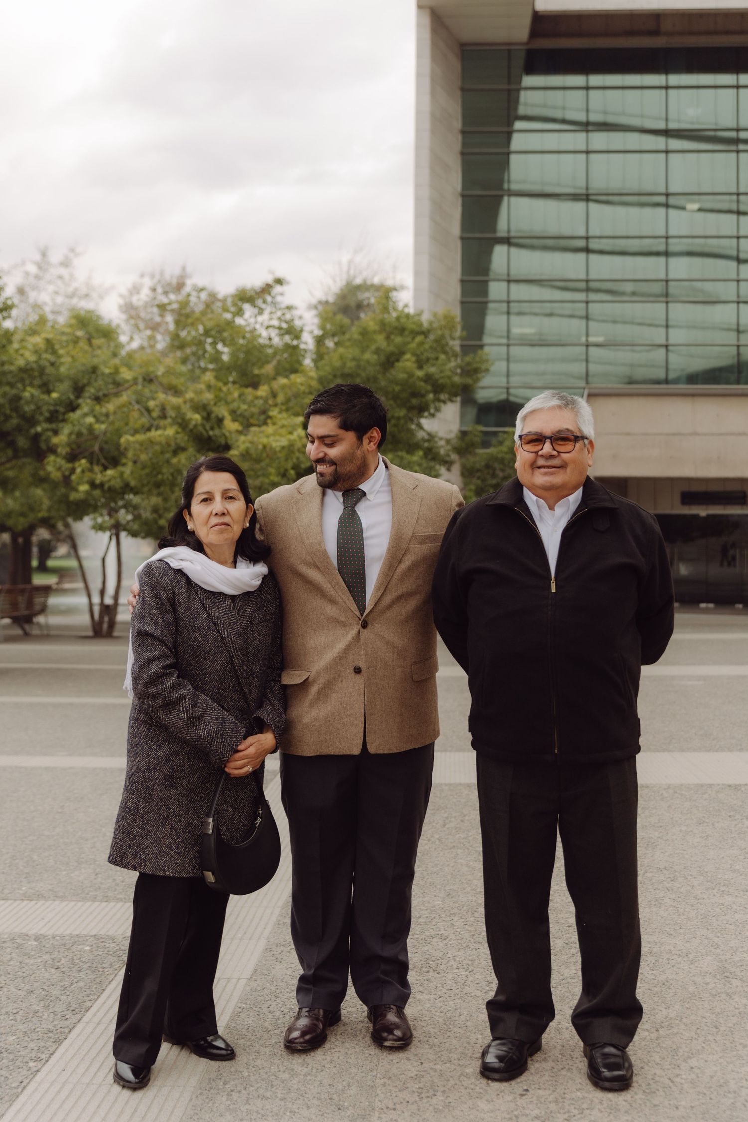 Groom in tan blazer posing with his parents outside a modern glass building on wedding day.