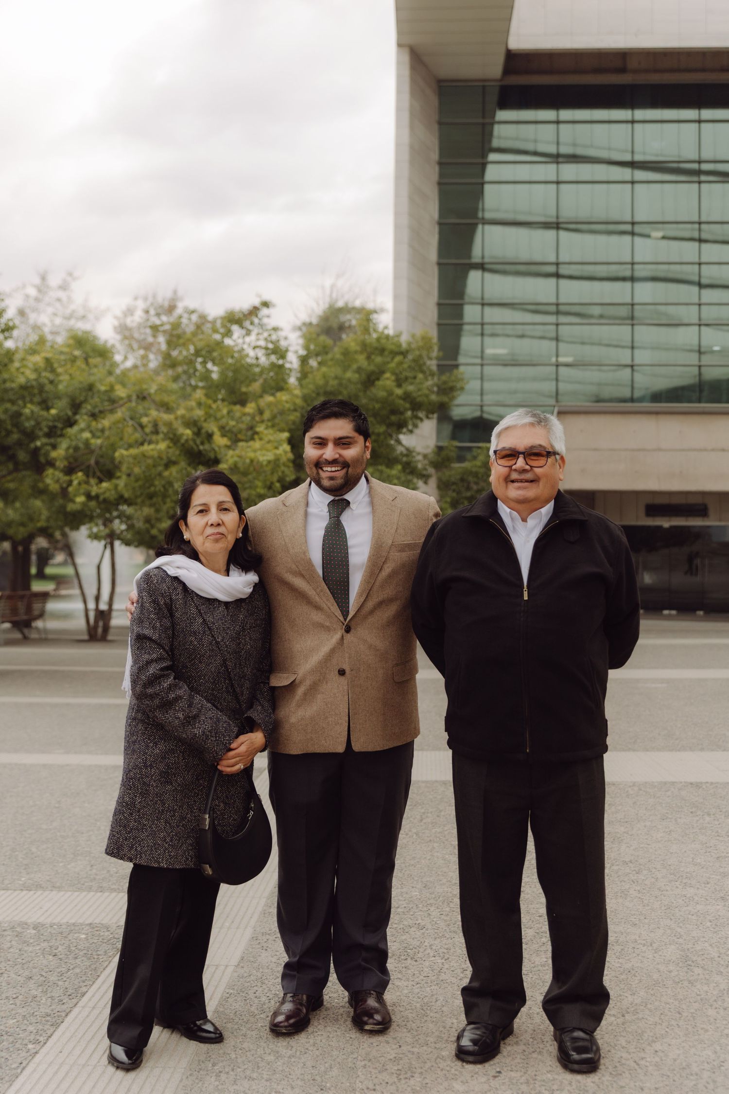 Groom in tan suit and tie standing proudly with parents outside modern building on wedding day.