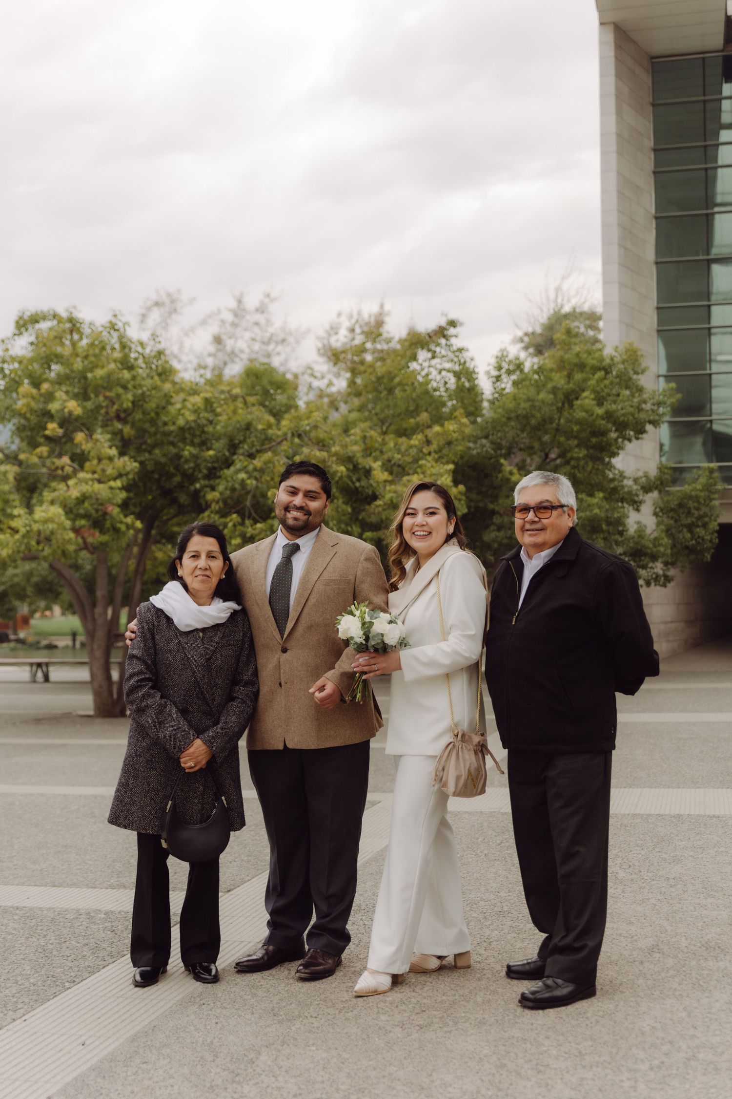 Newlywed couple in wedding attire posing with parents outside a modern building on their wedding day.