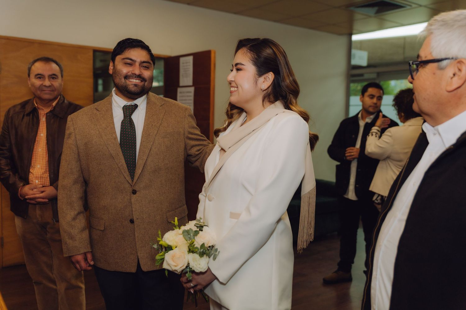Couple in formal attire holding white flowers greeting guests at civil wedding ceremony.