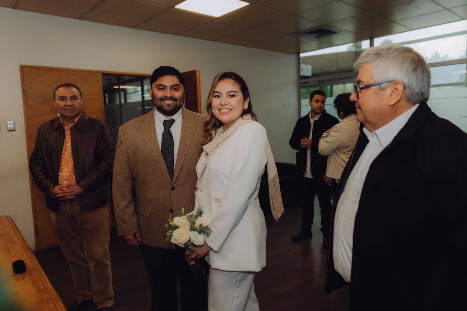 Bride in white suit and groom in brown blazer posing with family at civil wedding.