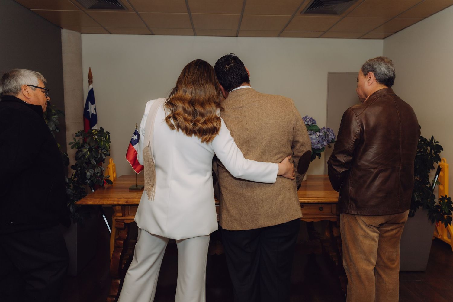 Newlywed couple standing together at wooden table with Chilean flag after civil ceremony.