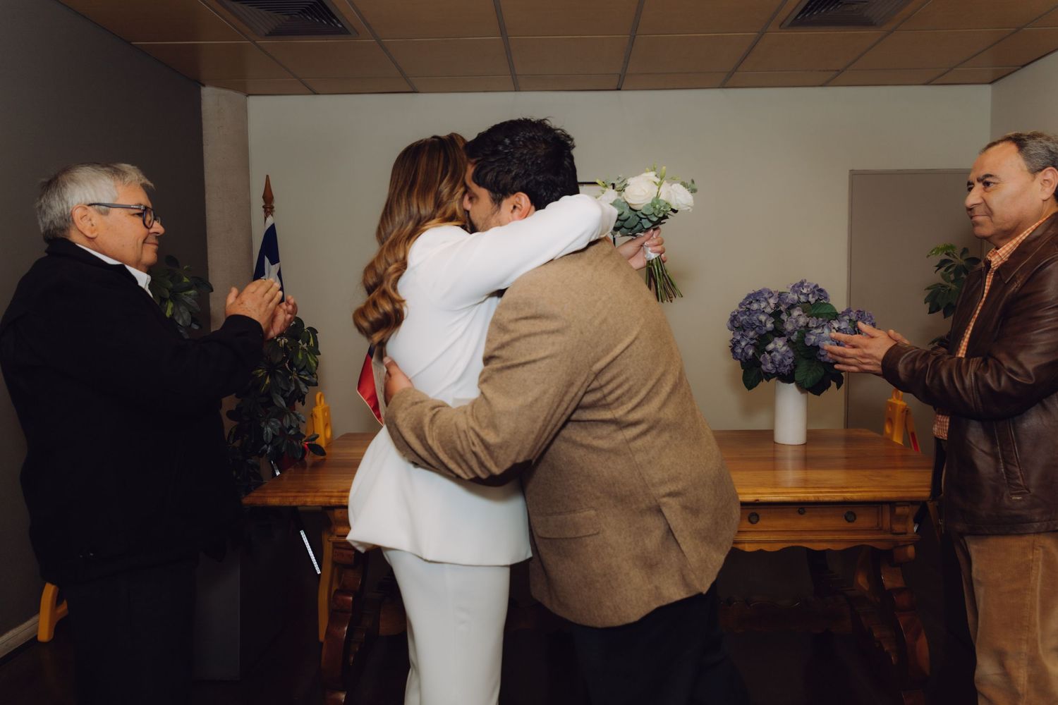 Couple sharing first kiss as newlyweds during intimate civil wedding ceremony with witnesses applauding.