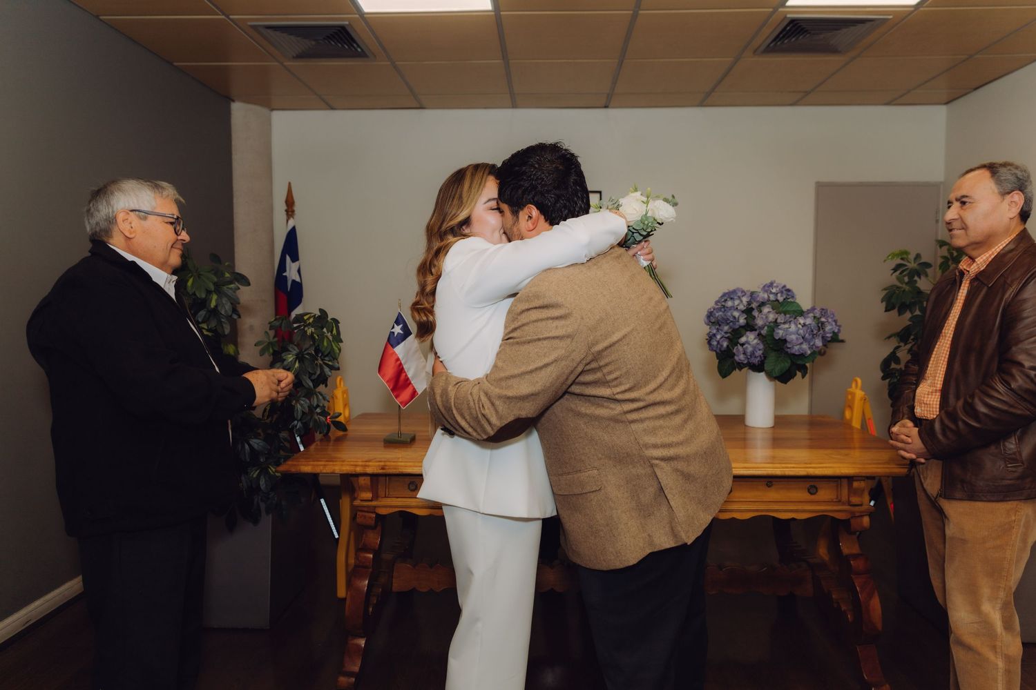 Bride and groom embrace warmly during civil wedding ceremony with Chilean flag and witnesses present.
