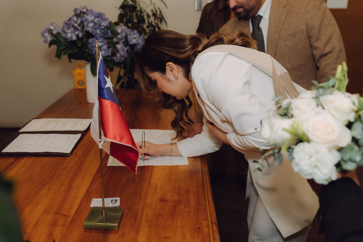 Bride in white suit signing official marriage documents at wooden table with Chilean flag during civil ceremony.