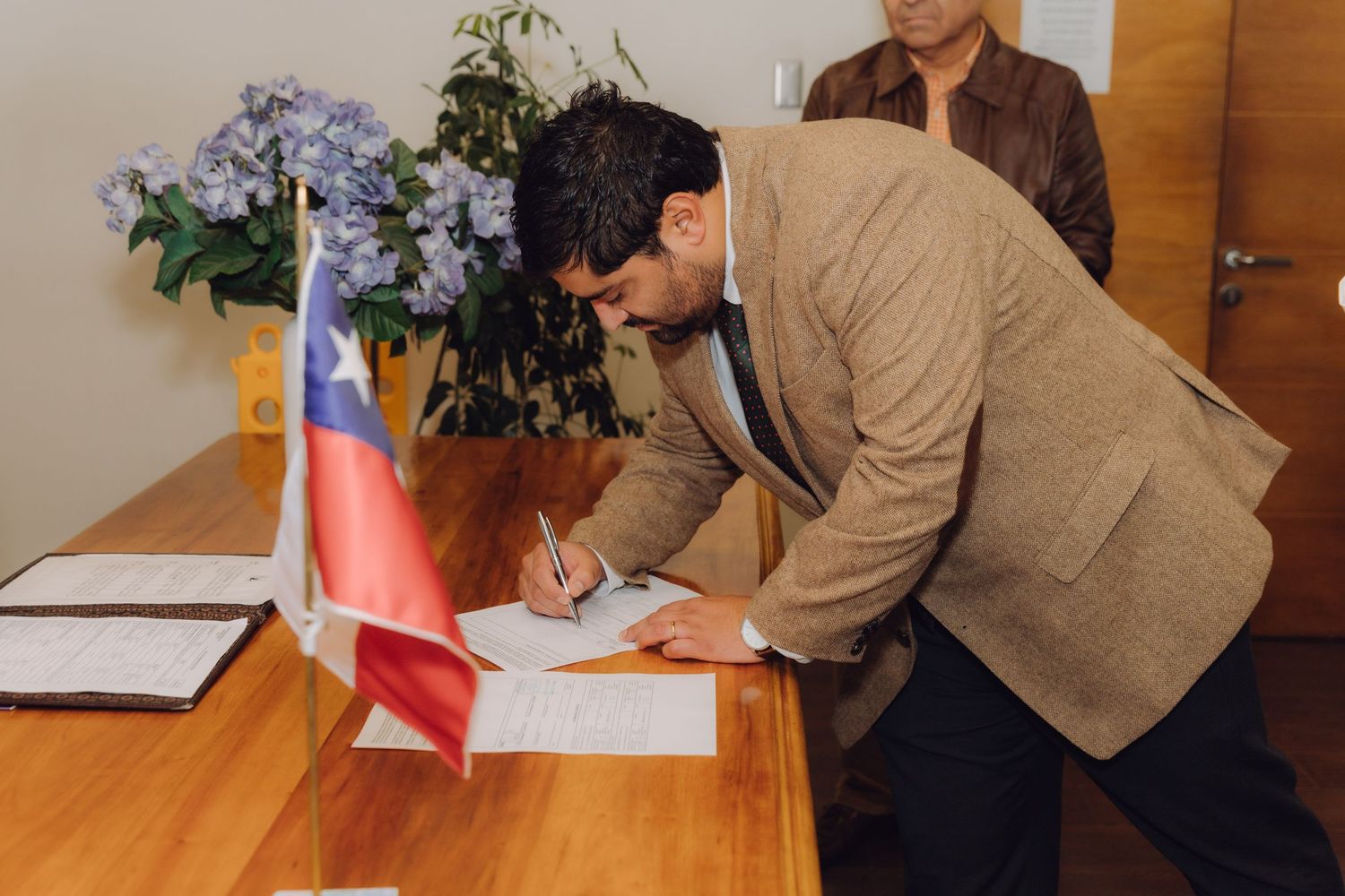 Groom in tan blazer signing marriage certificate at wooden table with Chilean flag during civil wedding.