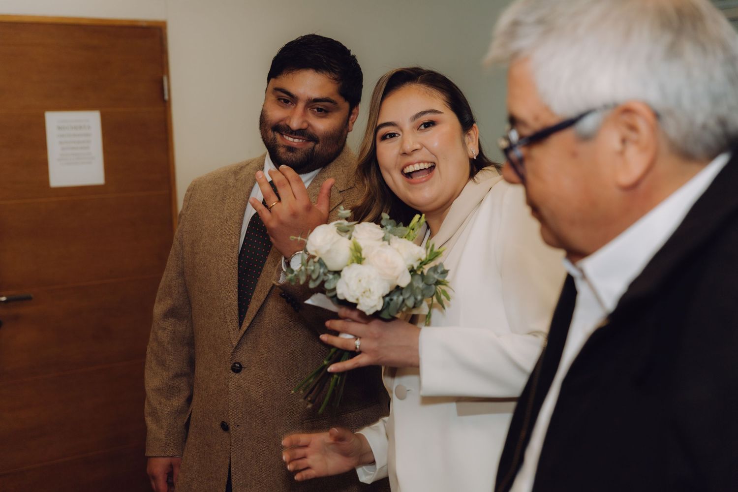 Joyful newlyweds smiling and showing wedding rings while holding white bouquet after civil ceremony.