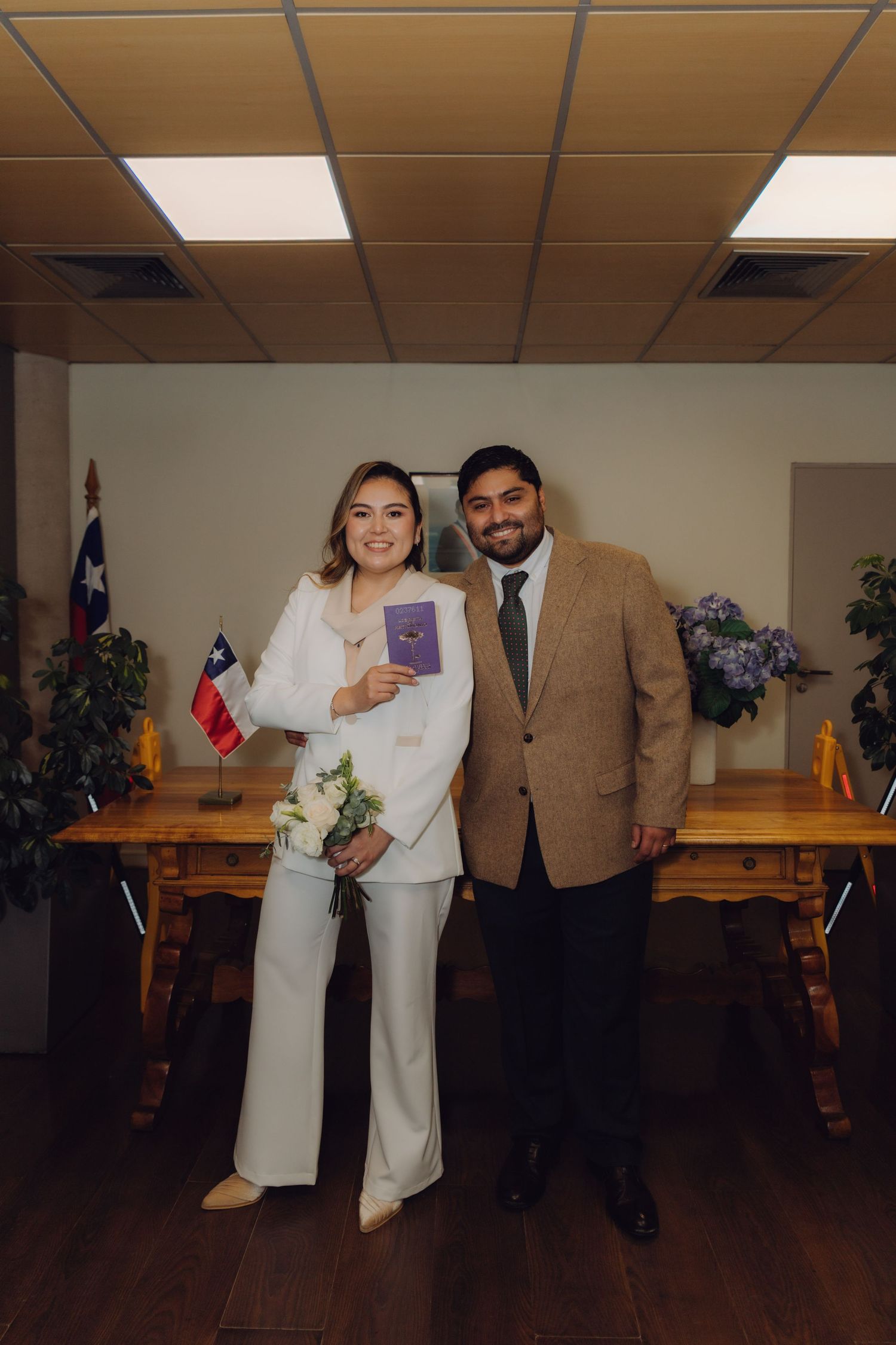 Couple in formal attire posing with marriage certificate and flowers in official registry office.