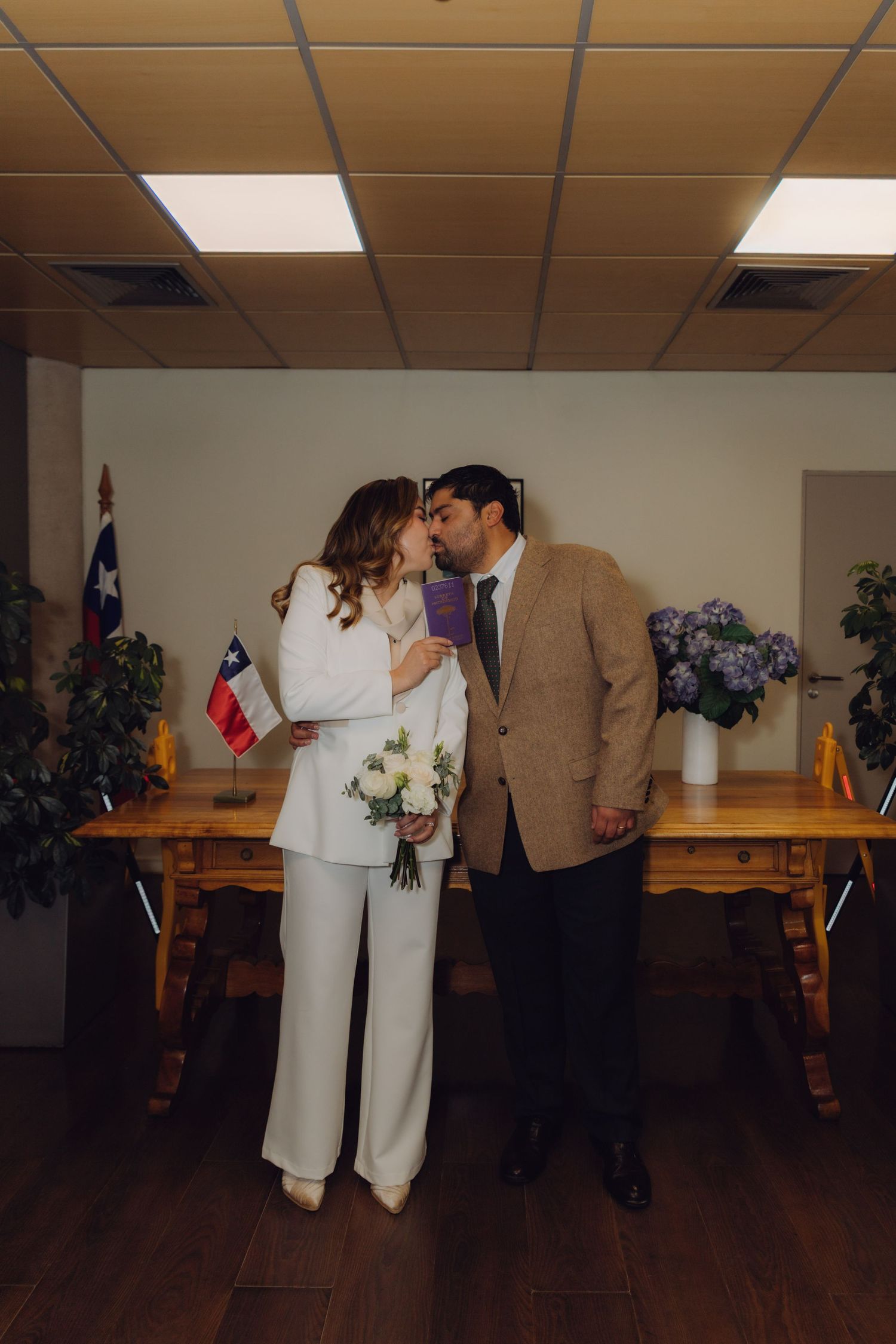 Newlyweds sharing a kiss while holding marriage certificate and white bouquet in civil registry office.