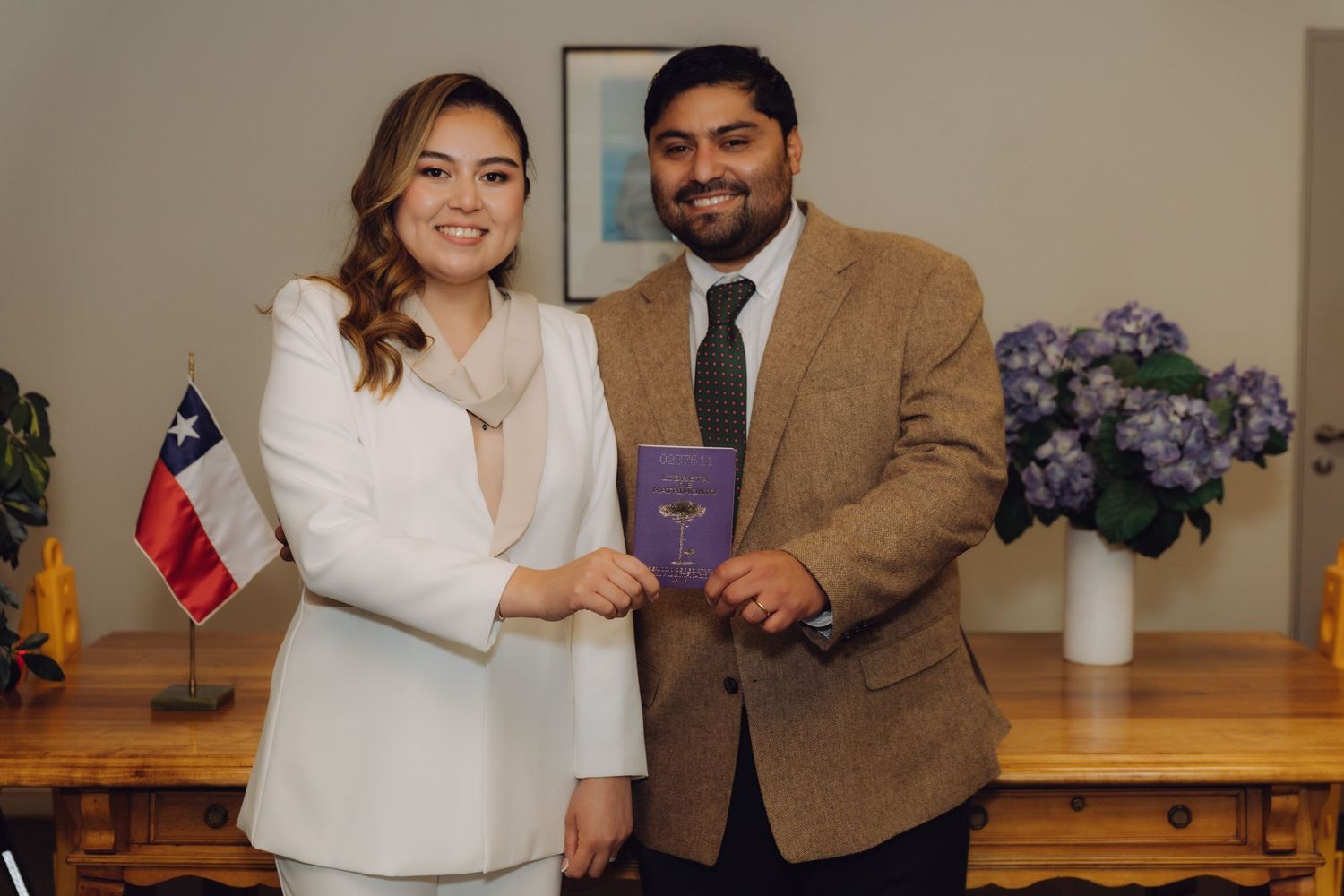 Smiling newlyweds posing together holding purple marriage certificate with Chilean flag in background.