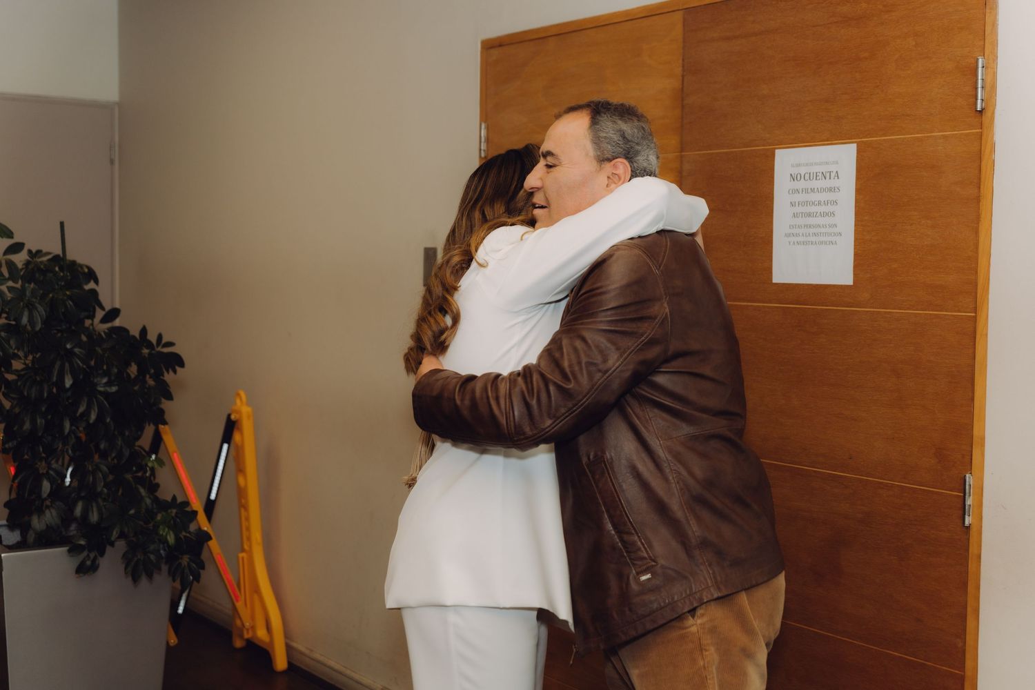 Bride in white suit sharing emotional hug with older man in brown leather jacket.