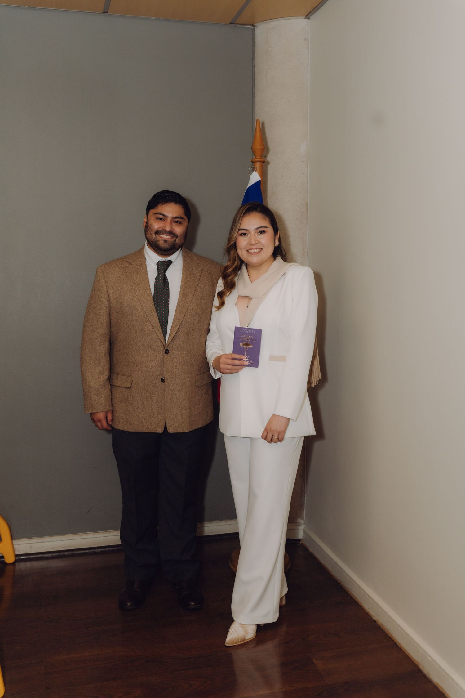 Couple posing beside Chilean flag, bride holding purple marriage booklet in registry office.