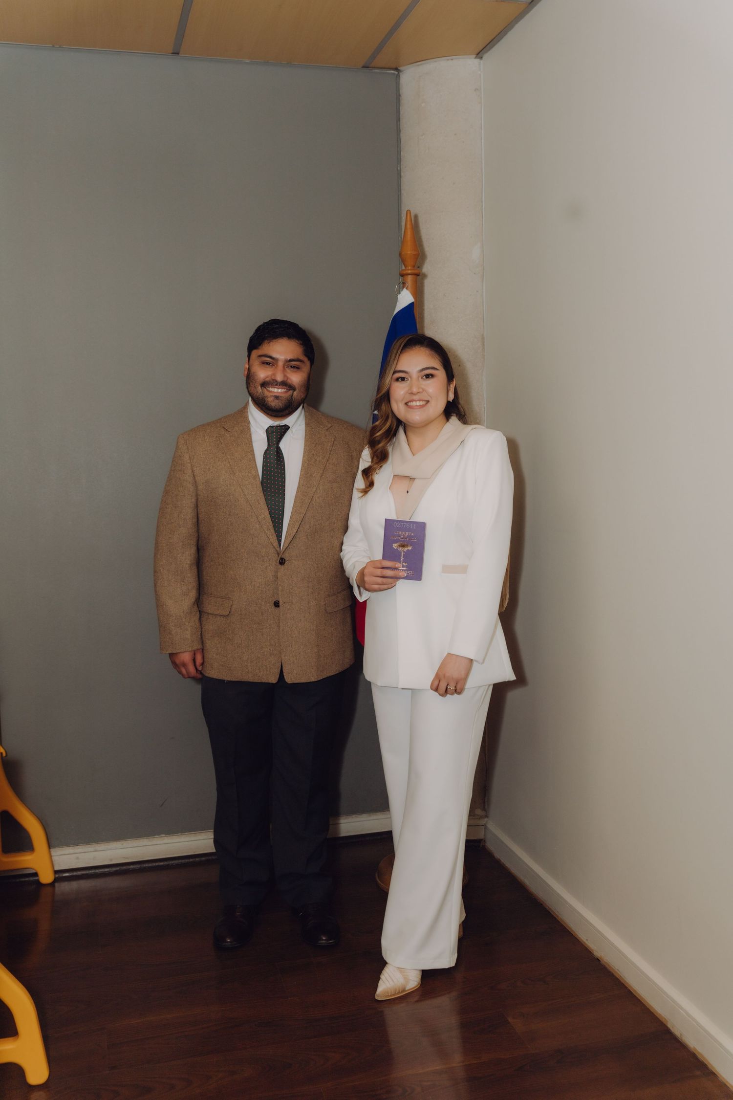 Smiling couple standing by Chilean flag, bride in white suit holding official marriage document.