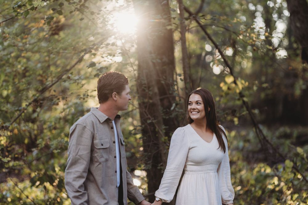 A couple in white and gray attire poses together in a sunlit forest setting during a romantic photoshoot.