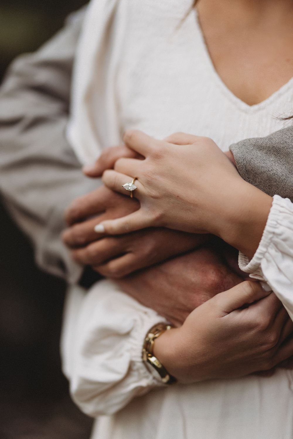Close up view of intertwined hands wearing gold wedding rings and white lace sleeves against a gray background.