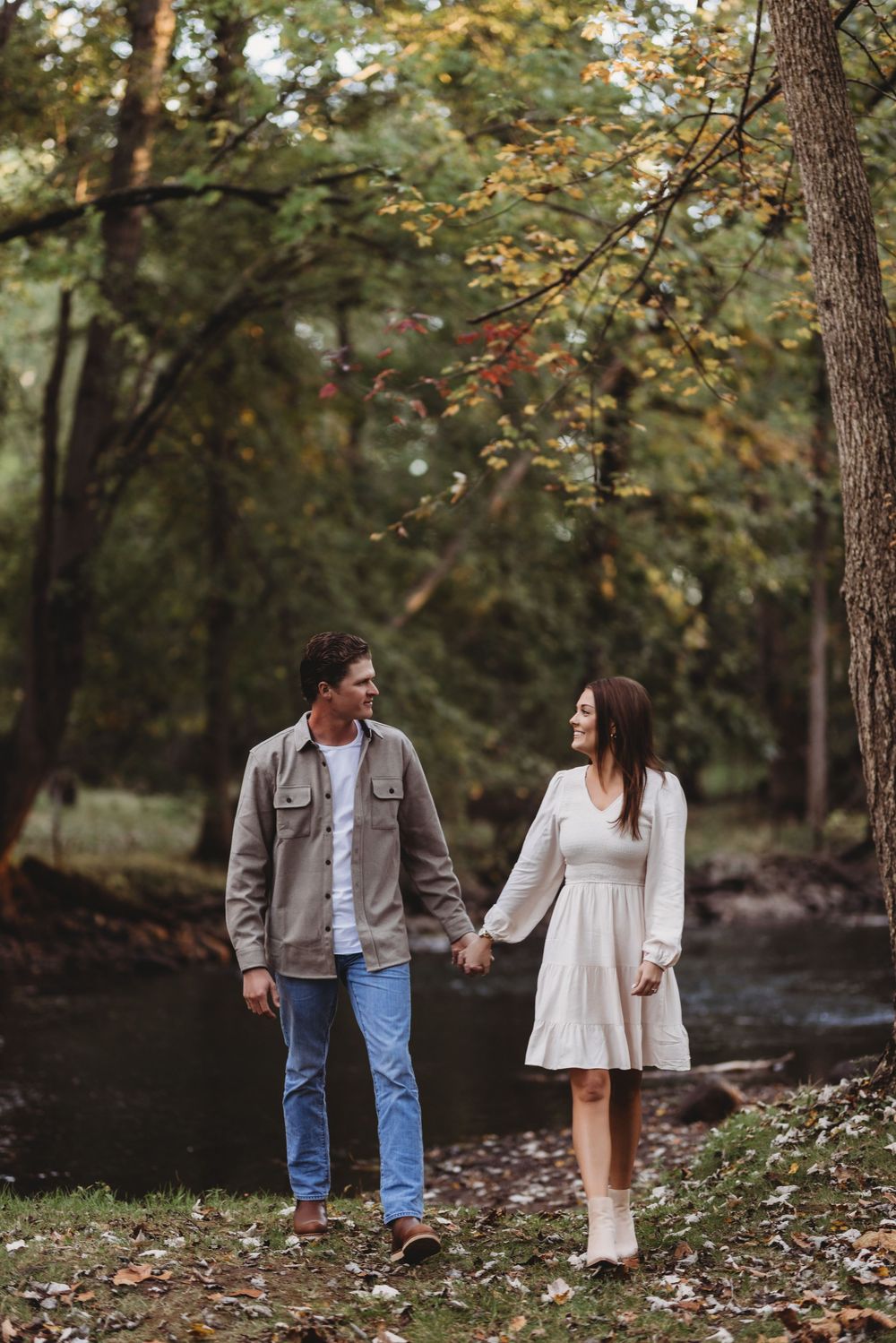 A romantic couple walks hand in hand along a wooded creek bank during autumn.