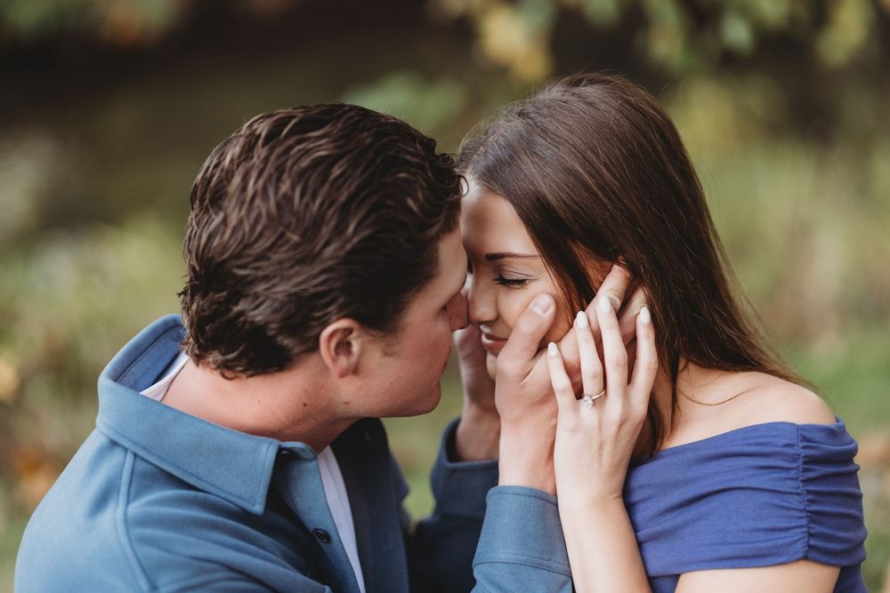 A romantic couple shares a tender kiss in a natural outdoor setting with soft lighting and greenery in the background.