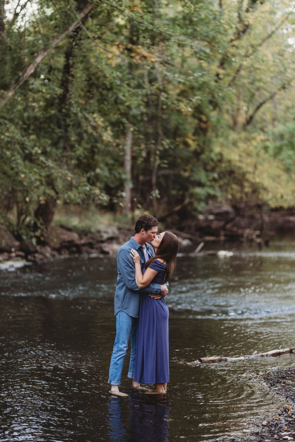 A couple embraces while standing in a flowing stream surrounded by lush forest scenery.