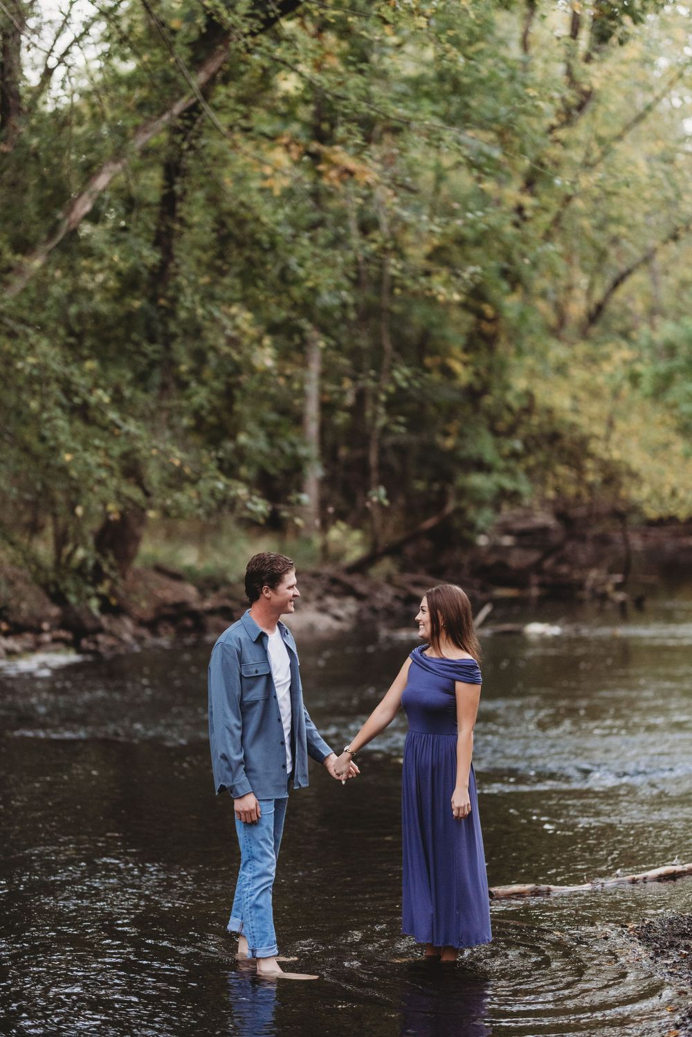 A couple stands barefoot in a shallow stream surrounded by lush forest.
