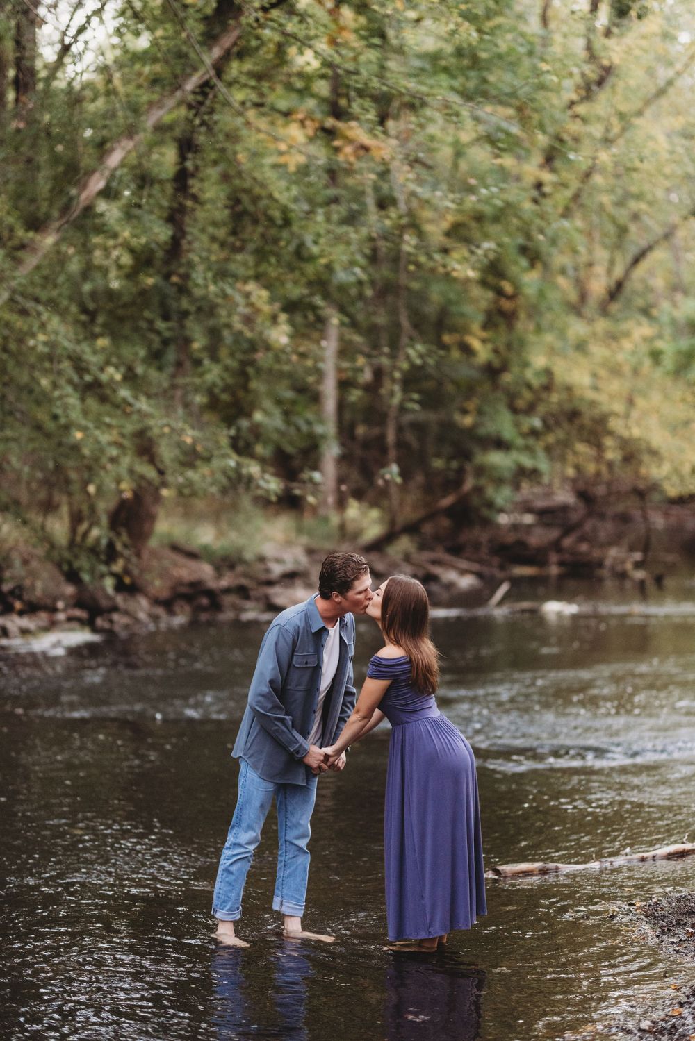 Couple stands together in shallow creek water surrounded by autumn trees.