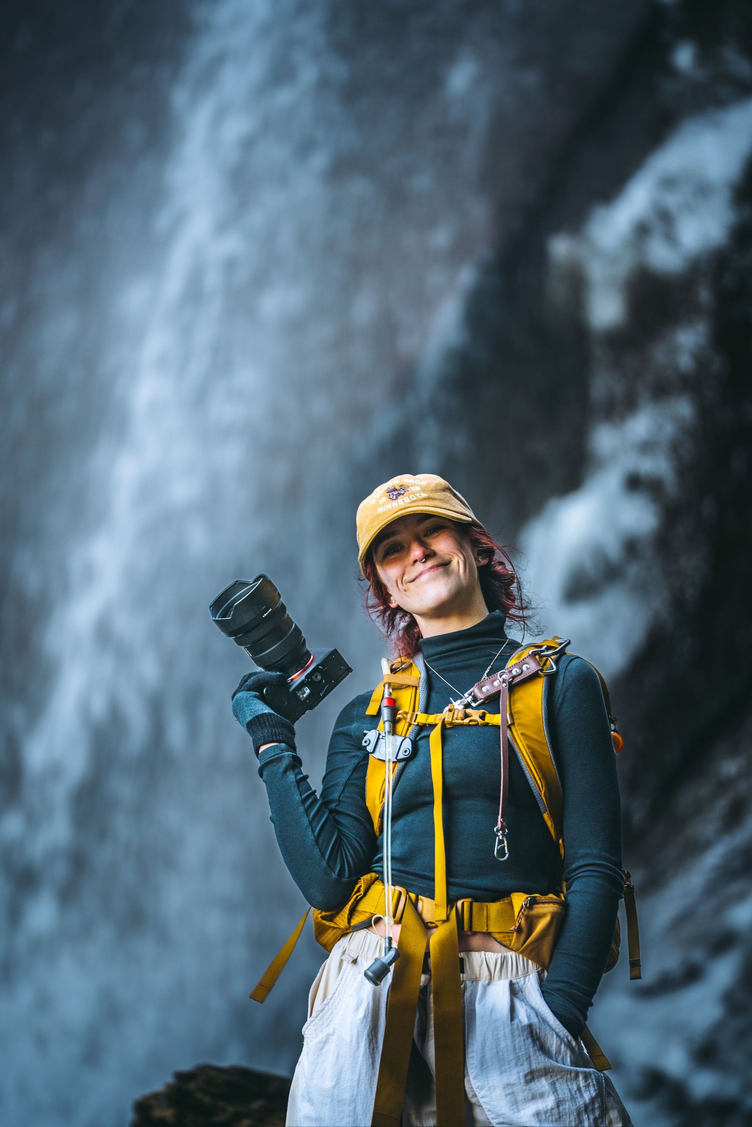 Female photographer with yellow backpack and cap smiling in front of a large waterfall.