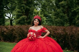 A stunning red quinceañera dress with floral embellishments photographed in a lush garden setting.