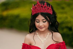 A stunning red ball gown with ornate crown and pearl necklace against a lush garden backdrop.