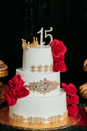 Elegant three-tiered white wedding cake decorated with gold filigree details and cascading red roses on a gold base.