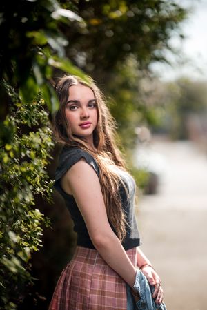 A person in a navy top and plaid skirt poses outdoors among green foliage with soft natural lighting.