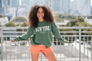 Someone wearing a green New York sweatshirt and orange shorts poses on an urban rooftop with city skyline in background.