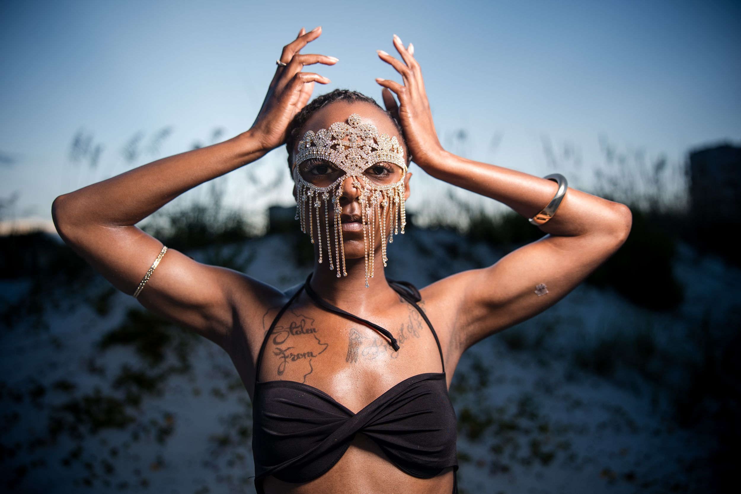 Person in black bikini top with tribal face paint and raised arms poses against dusky outdoor background.
