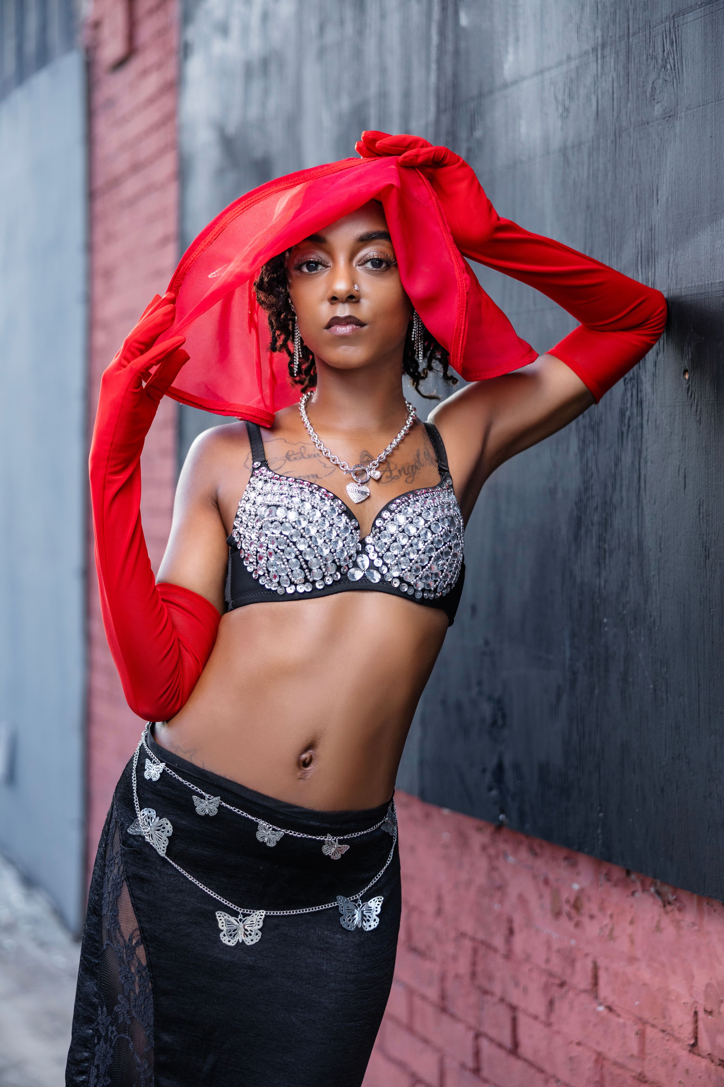 Fashion photo with red fabric flowing against pink and gray wall backdrop, featuring sparkly silver bra top and black pants.
