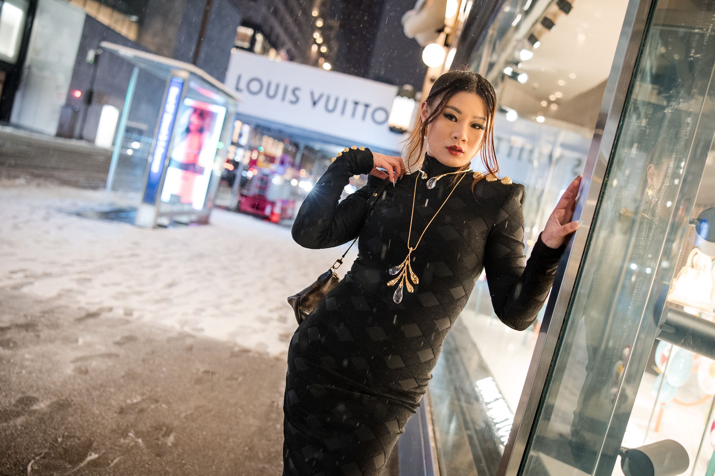 Person in black outfit poses in front of luxury retail stores at night with bright city lights in background.