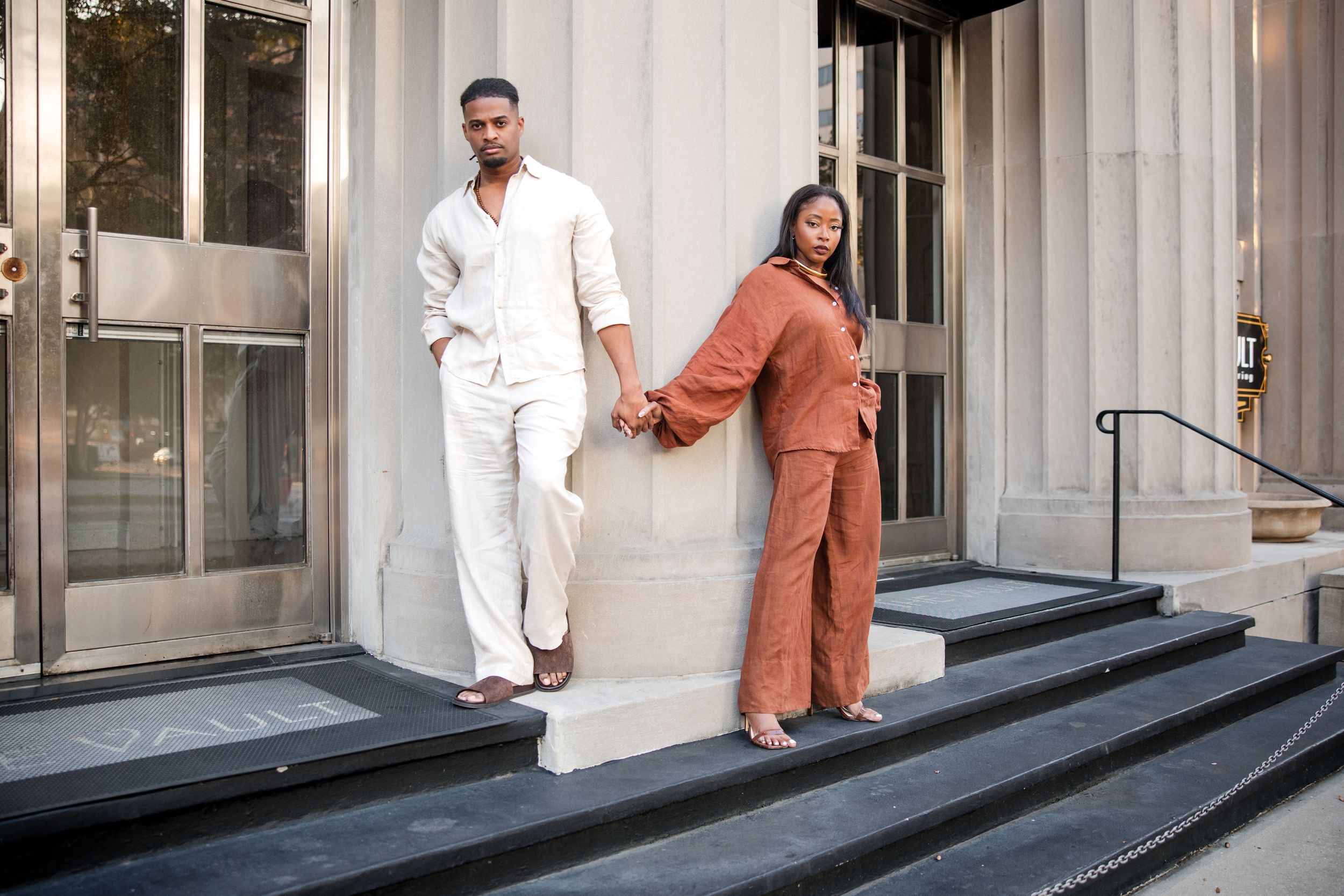 Two people in matching monochromatic outfits pose playfully on stone steps outside a building entrance.