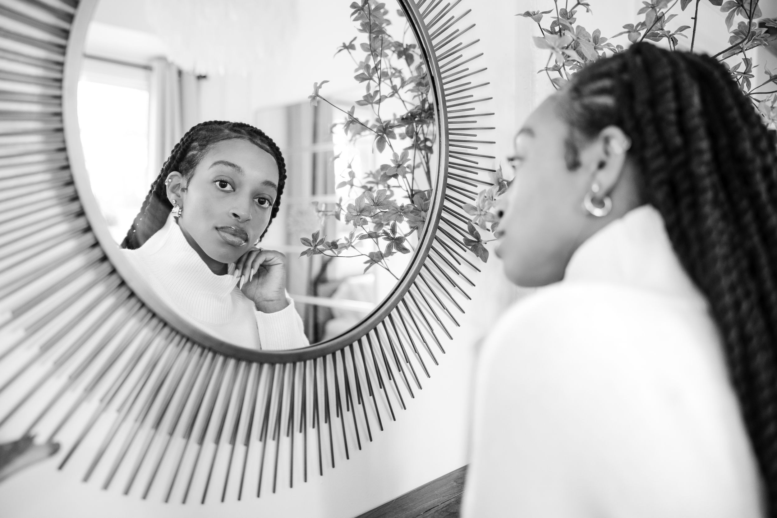 A reflection in a sunburst mirror shows someone with braided hair looking at their own reflection in black and white.