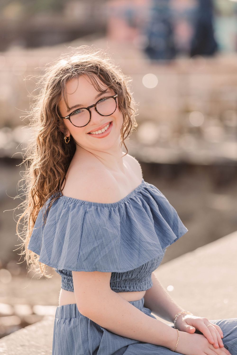 A portrait in a light blue off-shoulder summer dress with glasses and wavy hair against a blurred background.