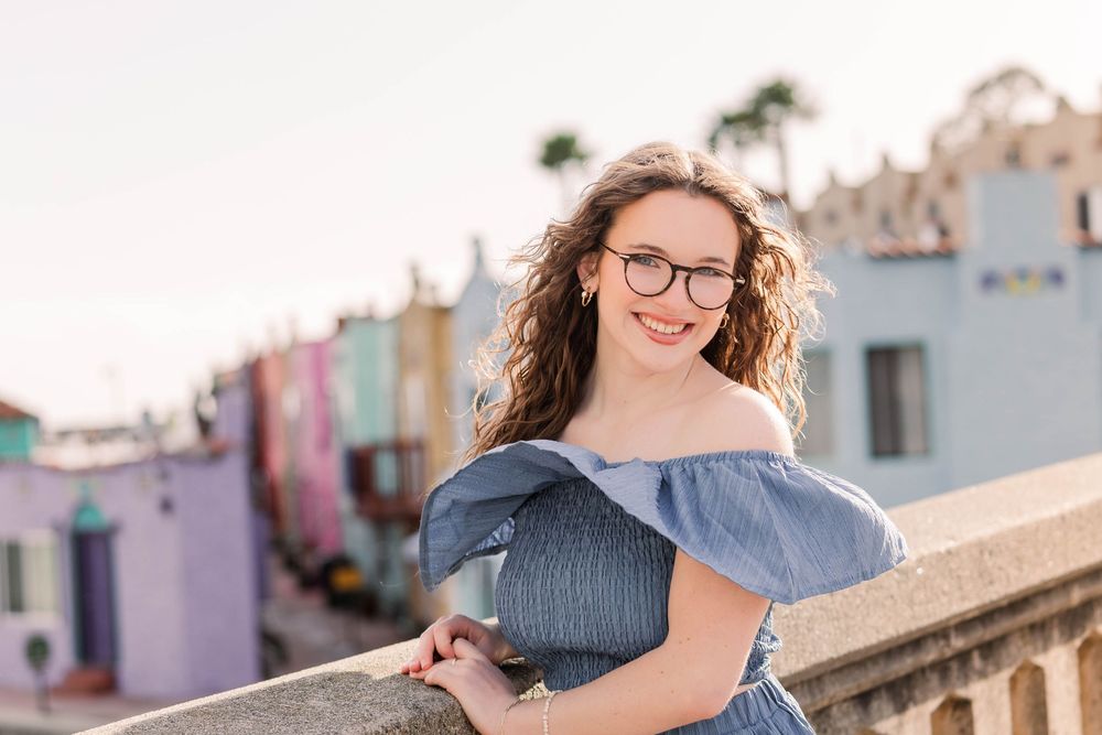 A person in a gray top sits on a stone wall with colorful pastel buildings in the background during sunset.