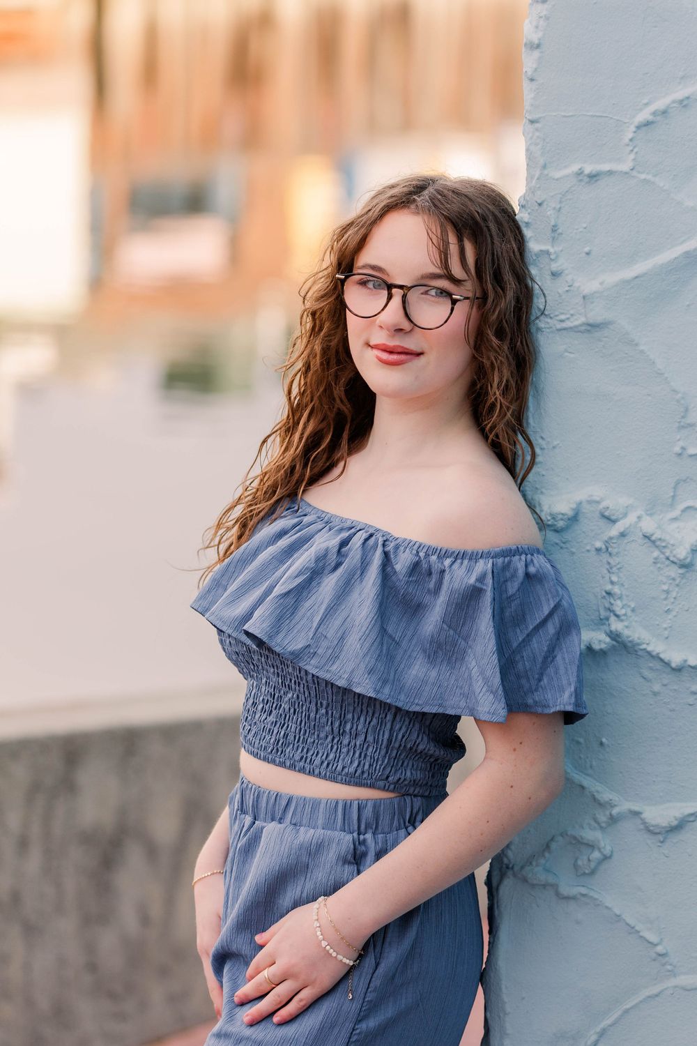 A portrait in front of a blue wall wearing glasses and a blue off-shoulder ruffle crop top outfit.