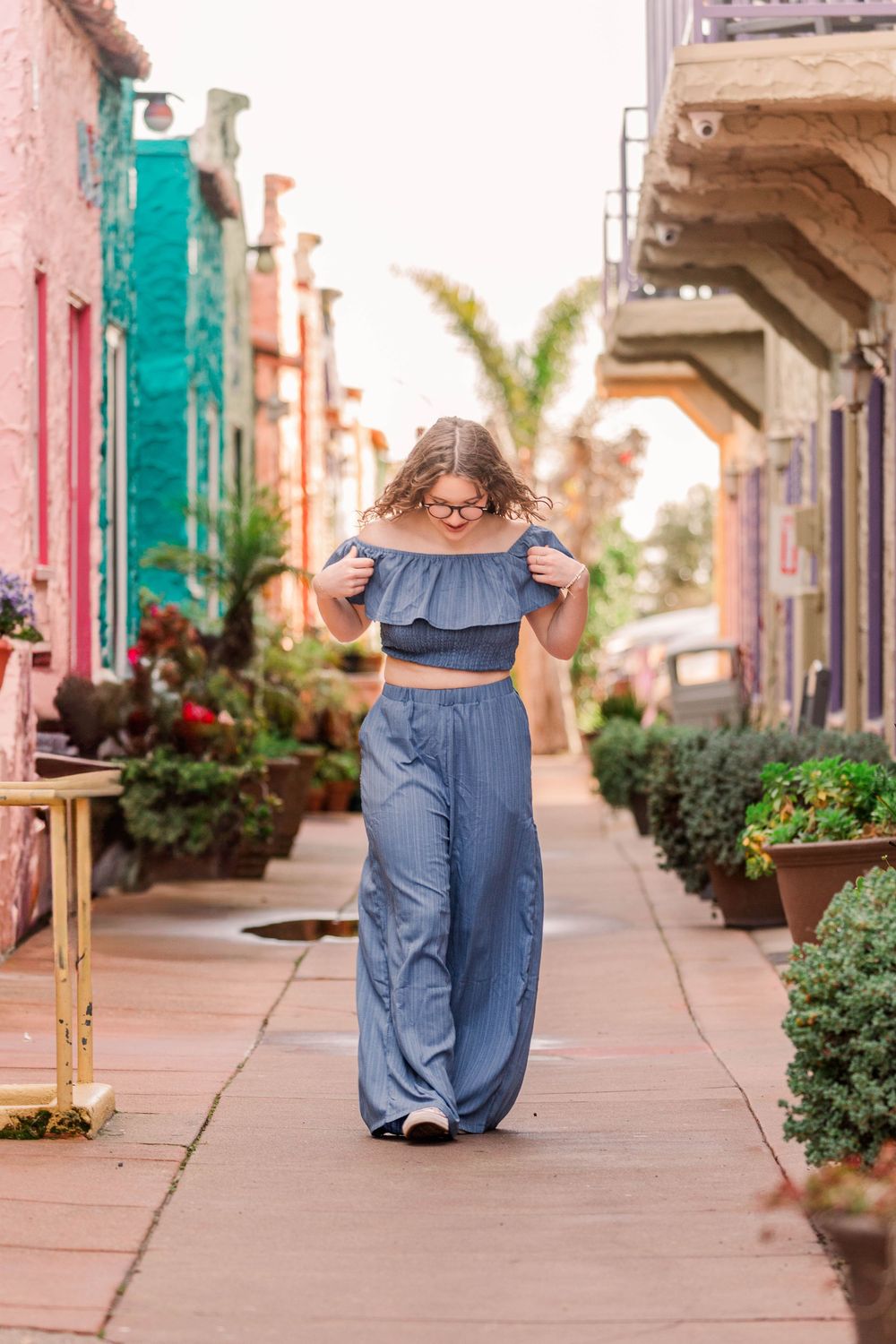 A toddler in a denim dress explores a colorful Spanish-style sidewalk lined with terracotta buildings and potted plants.