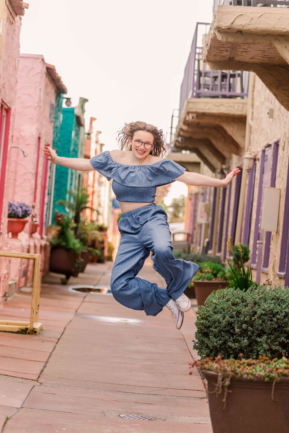 A person in denim performs a joyful jumping sequence on an urban sidewalk with pink buildings in the background.